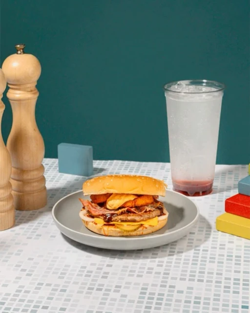 Burger served on gray plate with drink and salt and pepper shakers against teal background and white tiled surface.