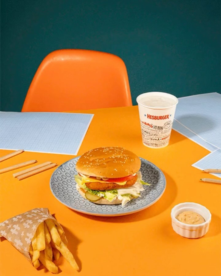Still life shot of a burger on a plate with fries, a drink, and a dipping sauce on an orange table against a green background.
