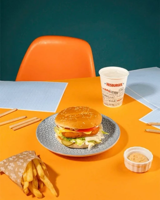 Still life shot of a burger on a plate with fries, a drink, and a dipping sauce on an orange table against a green background.