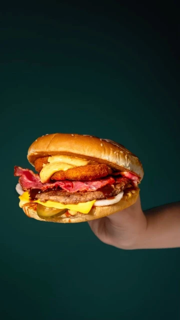 A hand holds a towering burger with beef, bacon, onion rings, cheese, and pickles, against a deep green background.