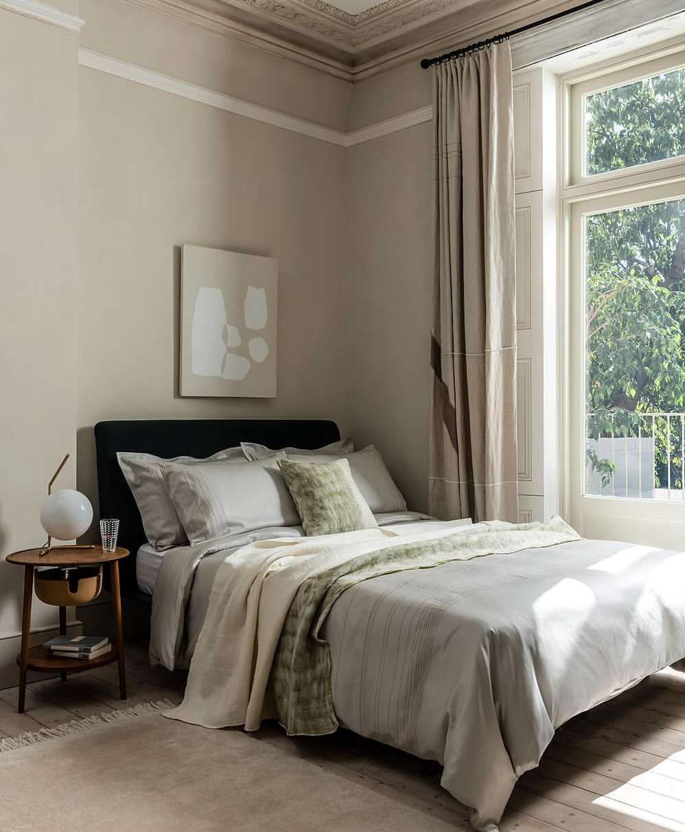 Bedroom interior featuring a made-up bed with neutral linens, a round side table with a lamp, and a large window with natural light.