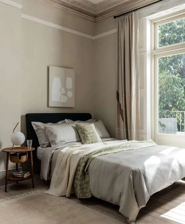 Bedroom interior featuring a made-up bed with neutral linens, a round side table with a lamp, and a large window with natural light.