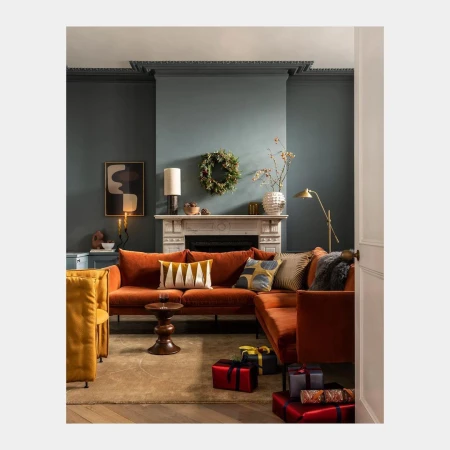 Cozy interior features an orange sectional sofa with decorative pillows, a fireplace with a wreath, and wrapped gifts.