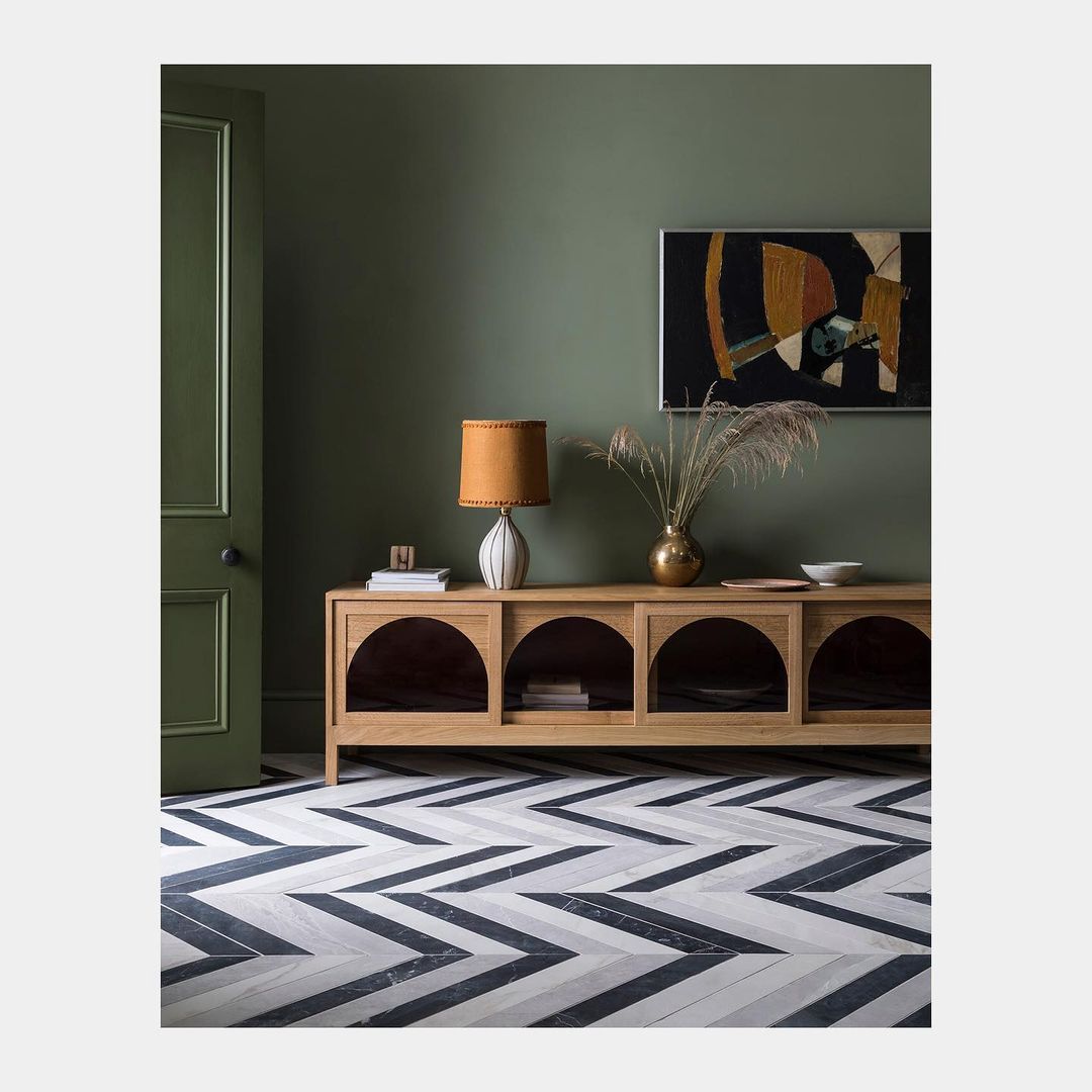 Living room interior with olive green walls, a light wood console table, and herringbone patterned floor, creating a modern, stylish space.