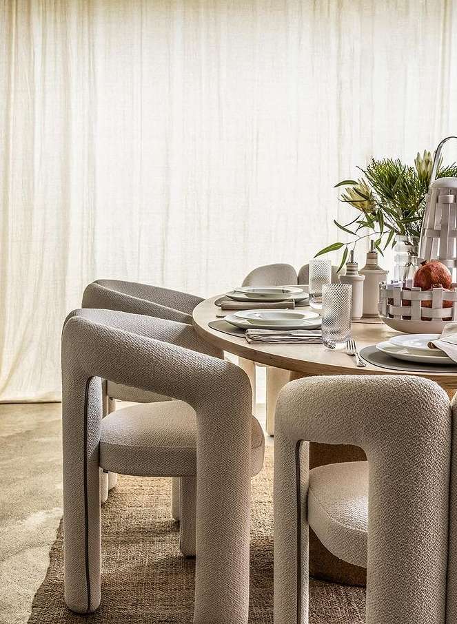 Round wood dining table set with upholstered modern chairs and neutral dishware in a minimalist dining room with natural light.