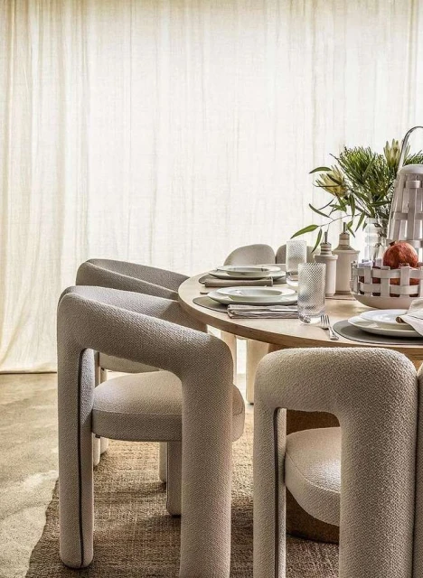 Round wood dining table set with upholstered modern chairs and neutral dishware in a minimalist dining room with natural light.