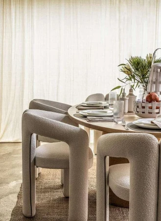 Round wood dining table set with upholstered modern chairs and neutral dishware in a minimalist dining room with natural light.
