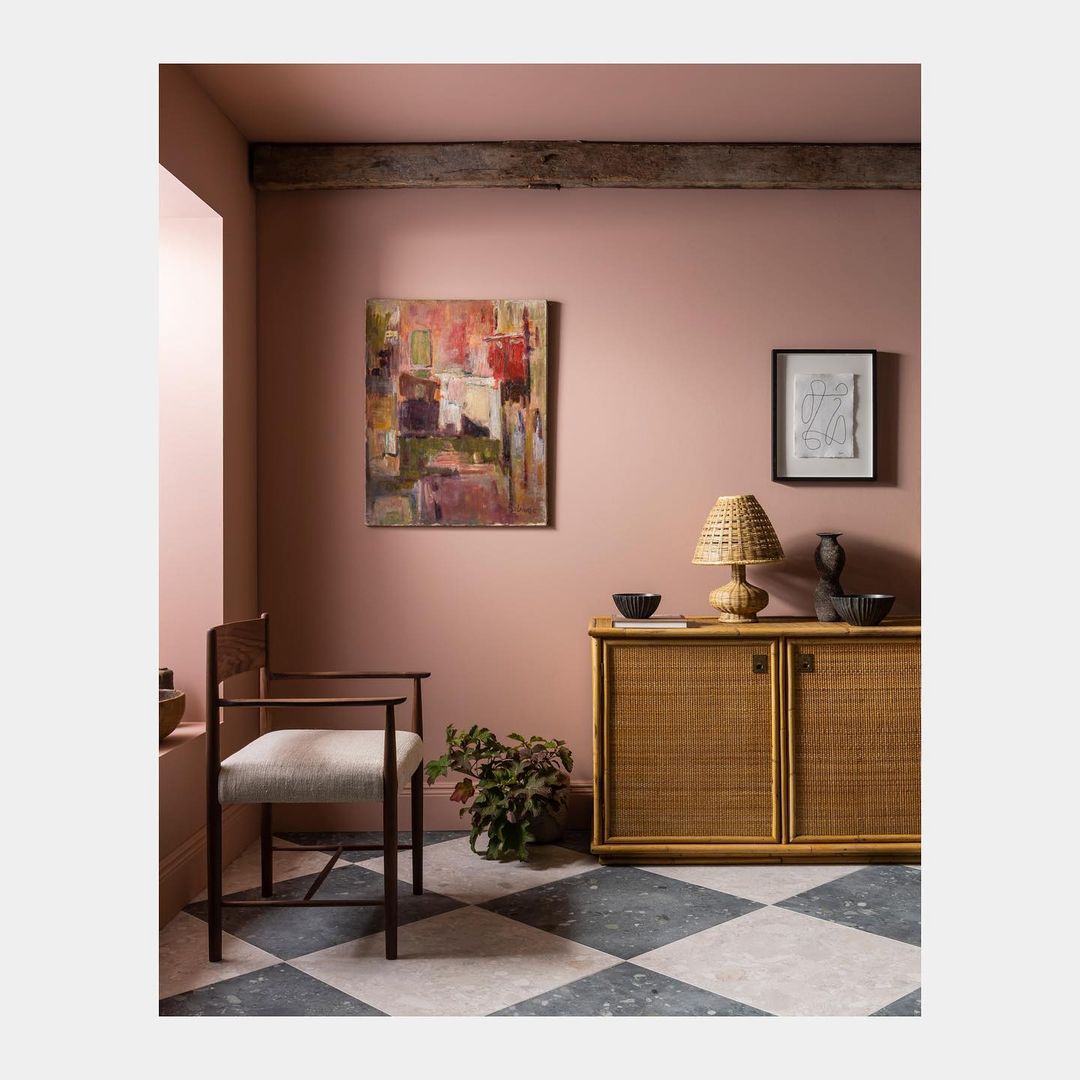 Interior design featuring a wooden chair, abstract art, a cabinet with a lamp, and a black and white checkered floor against a pink wall.