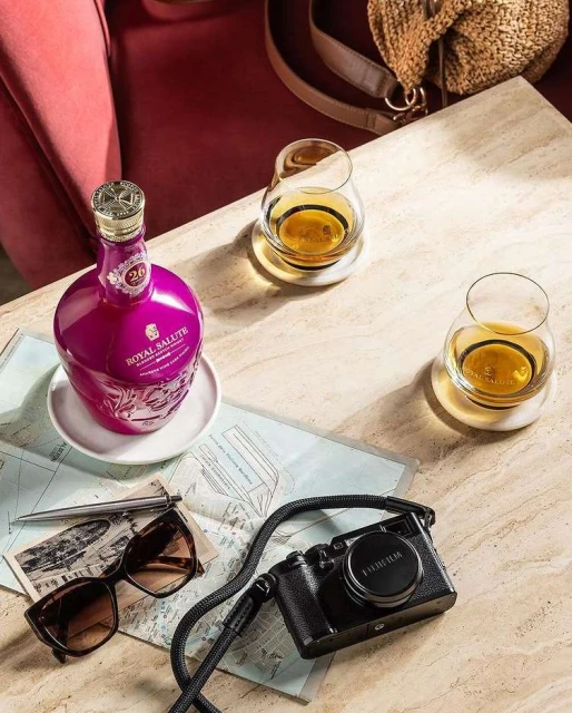 Still life shot of a purple Royal Salute whiskey bottle, two glasses, camera, and sunglasses on a table with a map underneath.