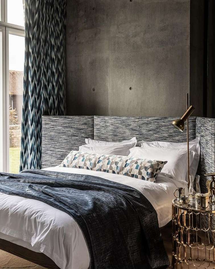Bedroom interior featuring a made bed with white linens and a blue blanket, a patterned headboard, and a concrete accent wall.