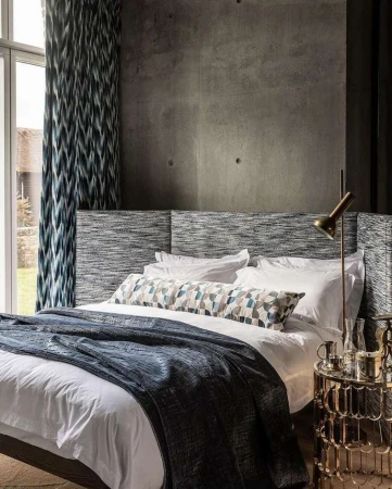 Bedroom interior featuring a made bed with white linens and a blue blanket, a patterned headboard, and a concrete accent wall.