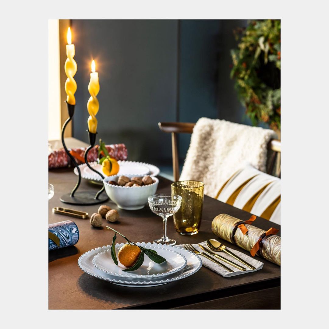 Table setting featuring lit candles, oranges, walnuts, glassware, white plates, and gold flatware create a festive and warm atmosphere.