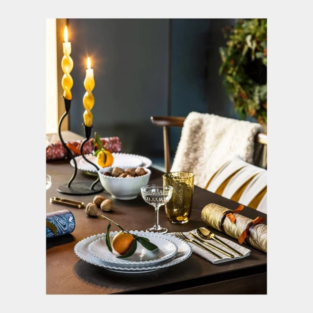 Table setting featuring lit candles, oranges, walnuts, glassware, white plates, and gold flatware create a festive and warm atmosphere.