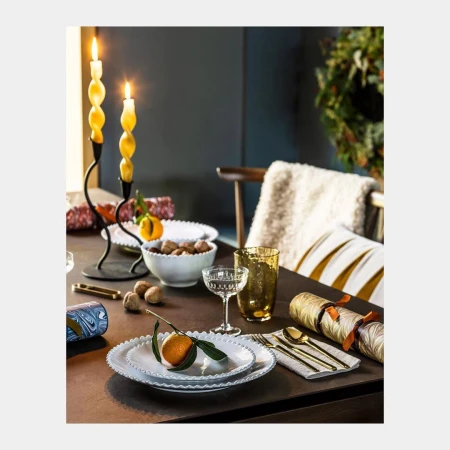 Table setting featuring lit candles, oranges, walnuts, glassware, white plates, and gold flatware create a festive and warm atmosphere.