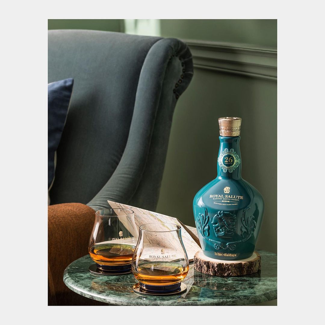 Turquoise Royal Salute whiskey bottle on a wood base with two glasses of whiskey on a green marble table next to a blue armchair.