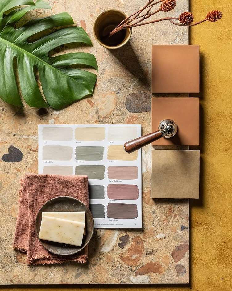 Still life shot with paint swatches, tiles, textiles, and tropical foliage, set on a terrazzo surface with a solid mustard background.