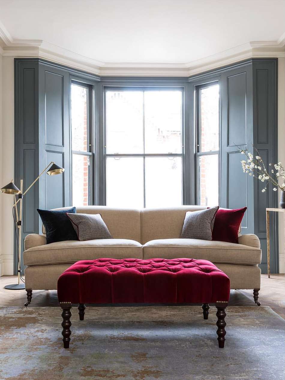 Living room interior featuring a beige sofa, a red velvet ottoman, pillows, a floor lamp, and a window with blue shutters.