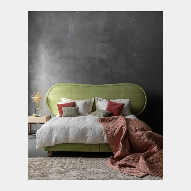 Interior with a green woven headboard bed, white bedding, patterned rug, and a dark grey textured wall in a residential setting.