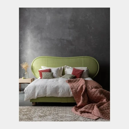 Interior with a green woven headboard bed, white bedding, patterned rug, and a dark grey textured wall in a residential setting.