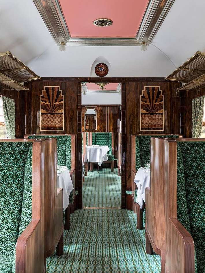 Elegant vintage train car interior featuring wood paneling, plush patterned seating, and formally set tables along a central aisle.