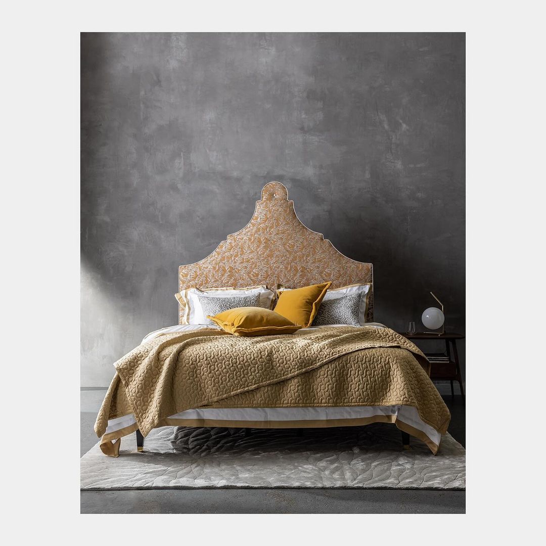 Bedroom with a decorative headboard, yellow quilt, pillows, and a gray textured wall in the background creates a cozy aesthetic.