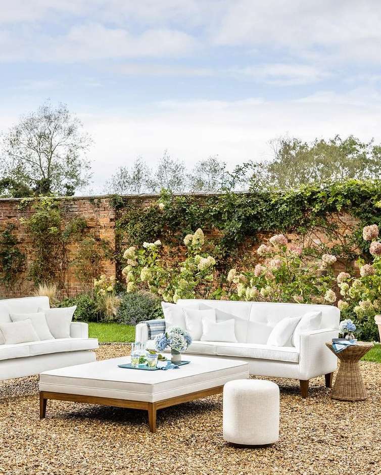 Outdoor furniture arrangement in a garden setting with white couches, ottoman, coffee table, and a small side table, all on a stone ground.