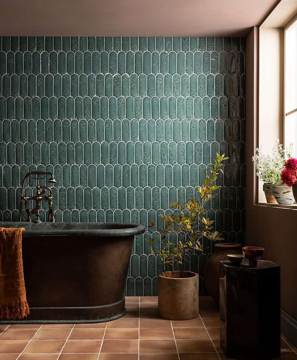 Bathroom interior with copper bathtub, green tiled wall, potted plants, and terracotta floor tiles in warm, inviting light.