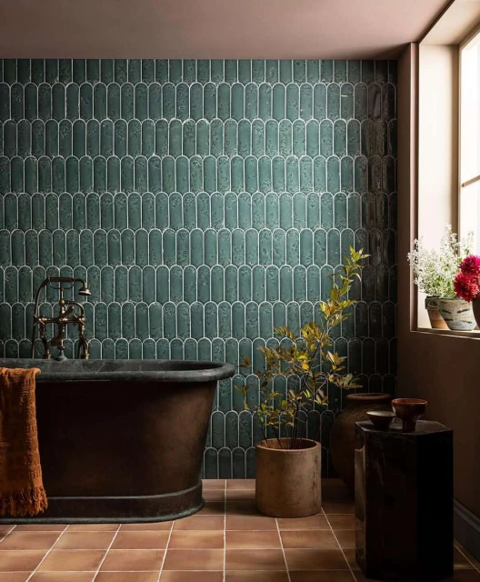 Bathroom interior with copper bathtub, green tiled wall, potted plants, and terracotta floor tiles in warm, inviting light.