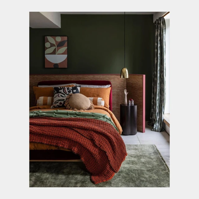 Bedroom interior with a made-up bed featuring a rust-colored knitted throw, patterned cushions, olive green walls, and a modern art print.