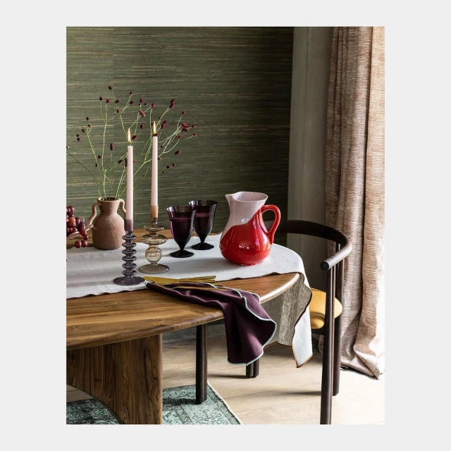 Still life on a round wooden table with candles, a vase, and a red pitcher, all set against a green wall and light-filtering curtains.