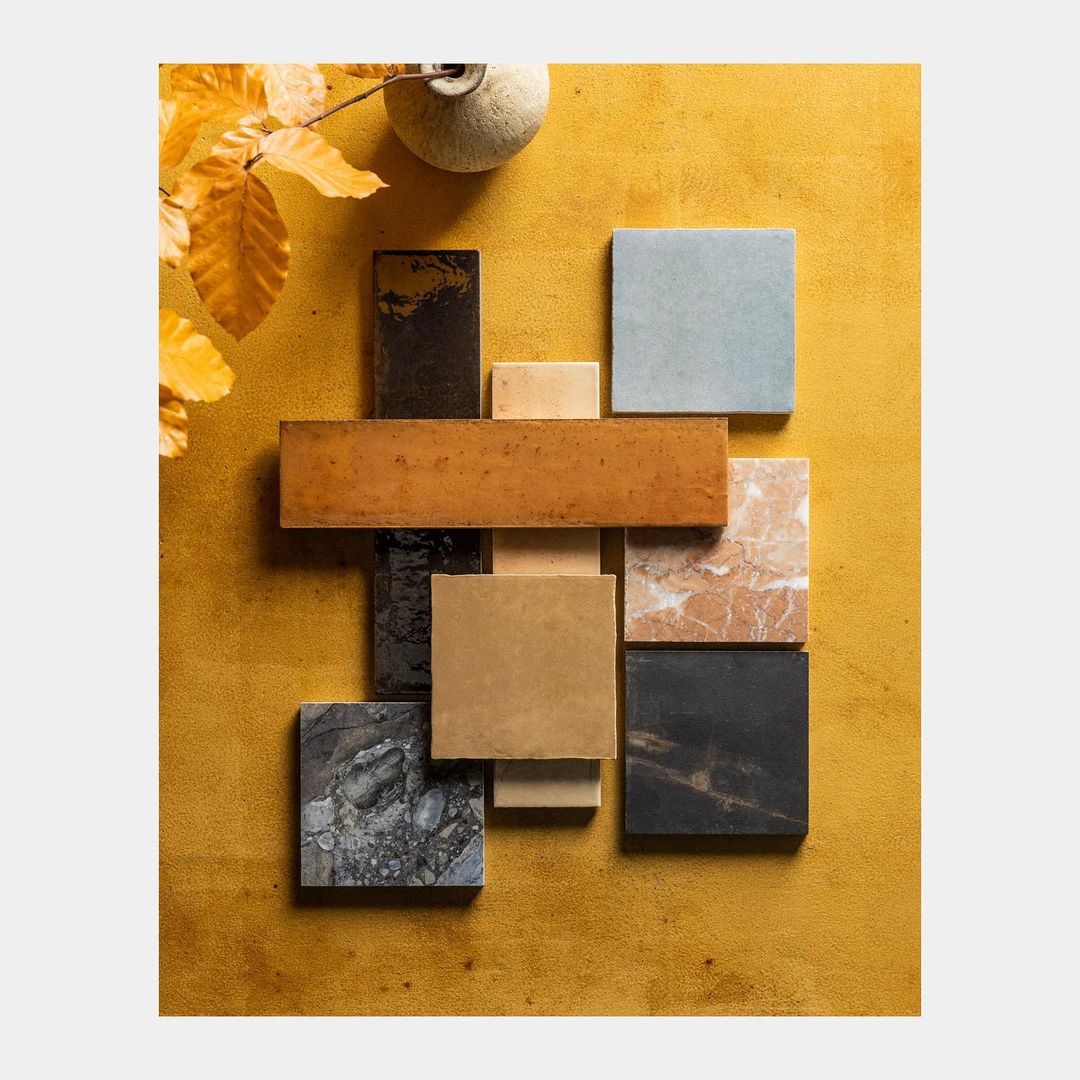 Still life of an arrangement of square and rectangular ceramic tiles in various colors on a yellow textured surface, with foliage.