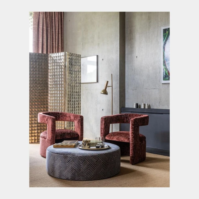 Interior with two modern armchairs, a patterned ottoman, and a brass floor lamp against a textured concrete wall in a living room setting.