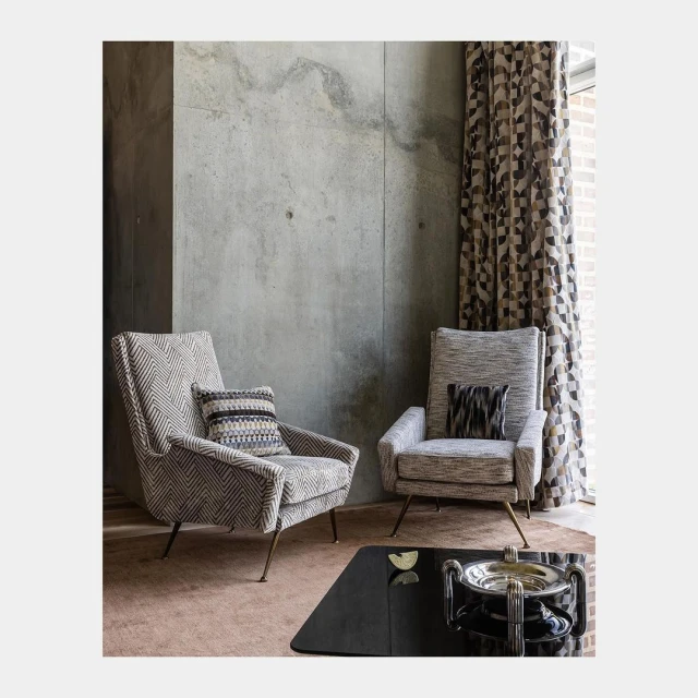 Interior design featuring two armchairs with patterned upholstery, decorative cushions, and a black coffee table against a textured grey wall.