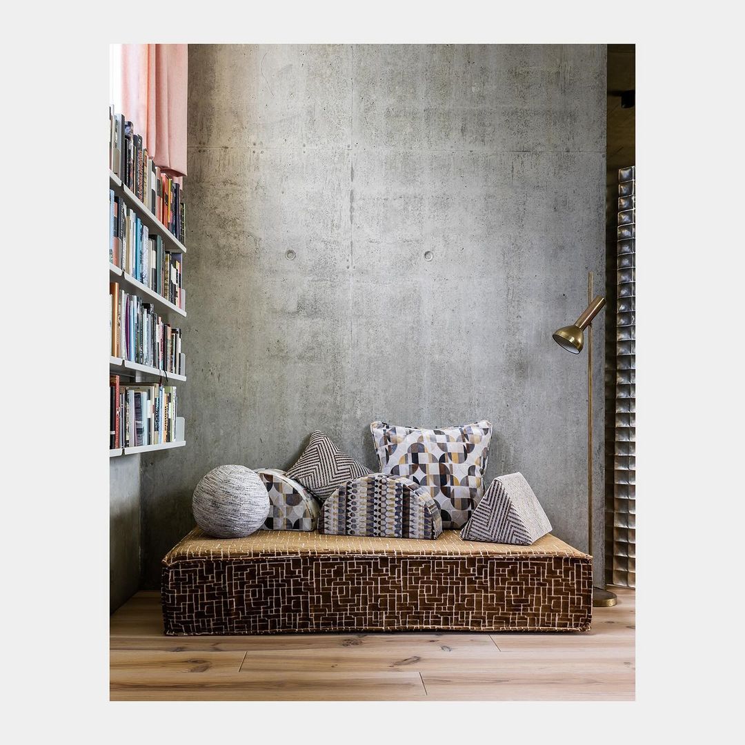Interior of a room features a daybed covered with decorative pillows, complemented by a bookshelf filled with books and a concrete wall.