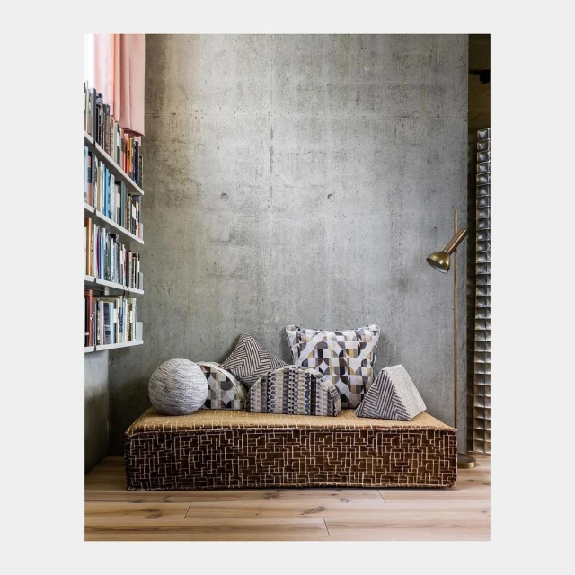 Interior of a room features a daybed covered with decorative pillows, complemented by a bookshelf filled with books and a concrete wall.