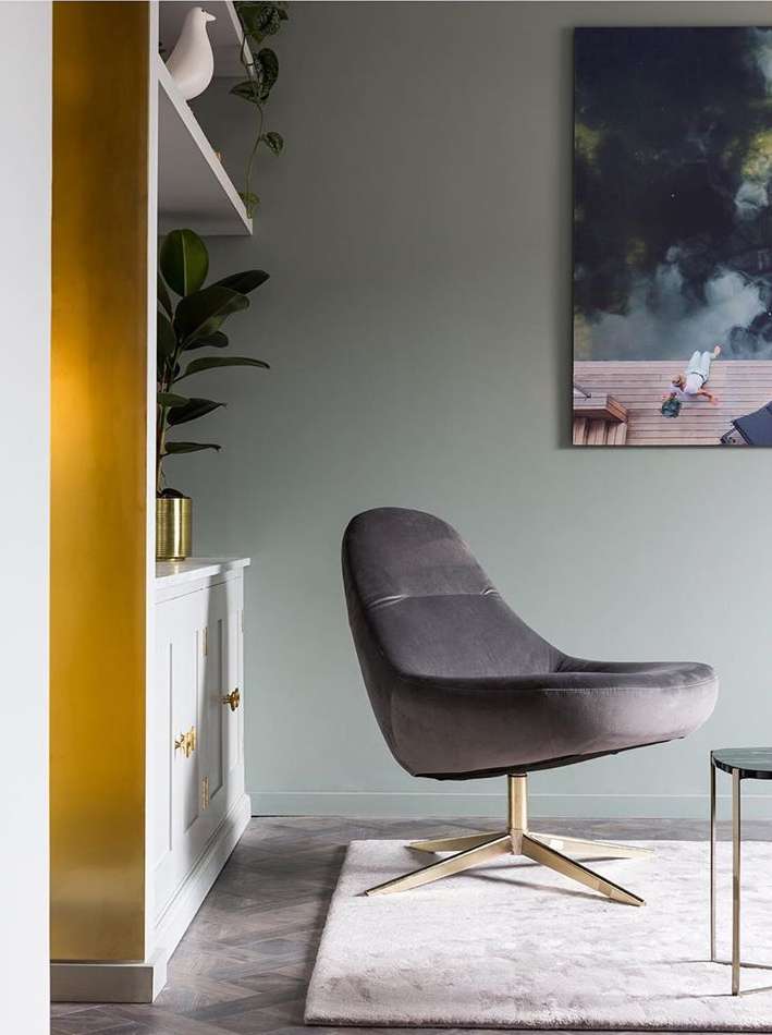 Interior shot of a grey swivel chair with gold base on a rug near a side table and white cabinet, with sage walls and modern art.