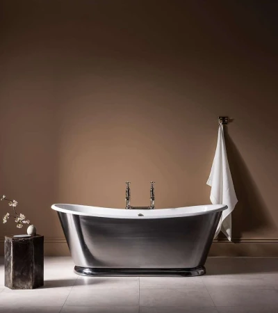 Freestanding metal bathtub in a warm brown room with a white towel hanging on the wall and a small vase on a pedestal.