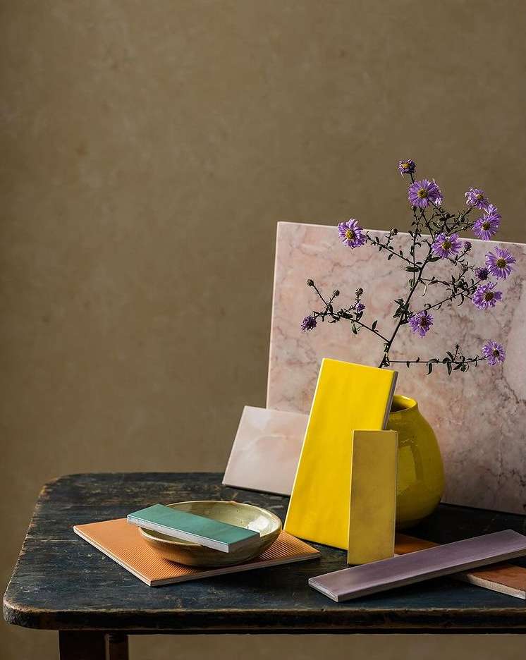 Still life on a blue wooden table features ceramic tiles in various colors, a yellow vase with purple flowers, and neutral wall paint.
