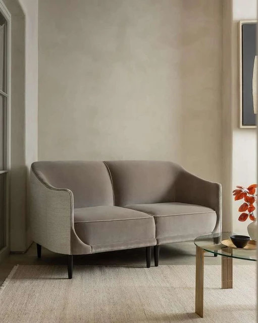 Interior shot of a neutral-toned living room featuring a modern loveseat sofa, glass table, textured rug, and minimalist wall art.