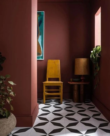 Yellow wooden chair against terracotta walls with geometric black and white tile flooring and a side table with lamp.