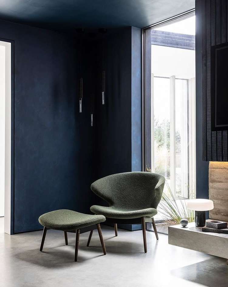 Modern living room interior featuring a green lounge chair with ottoman, dark blue walls, and natural light from a large window.