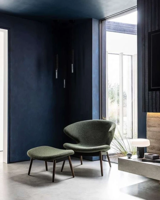 Modern living room interior featuring a green lounge chair with ottoman, dark blue walls, and natural light from a large window.