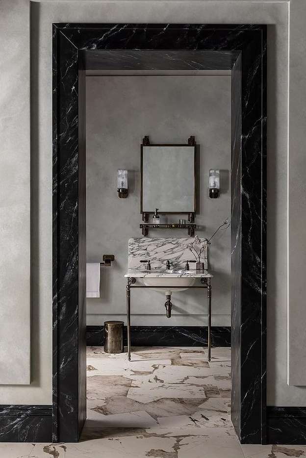 Bathroom interior with marble sink, mirror, sconces, and neutral wall and floor tiles, viewed through marble doorway.