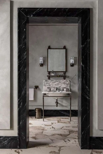 Bathroom interior with marble sink, mirror, sconces, and neutral wall and floor tiles, viewed through marble doorway.