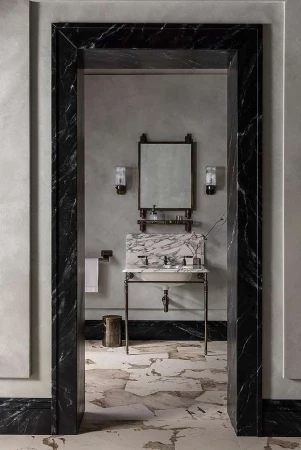 Bathroom interior with marble sink, mirror, sconces, and neutral wall and floor tiles, viewed through marble doorway.