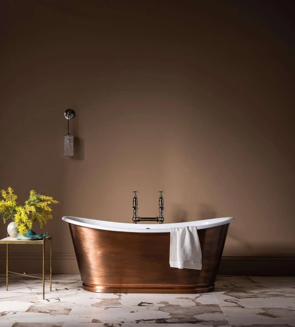 Elegant bathroom interior featuring a copper bathtub with white towel, marble floor, and a vase of yellow flowers on a side table.