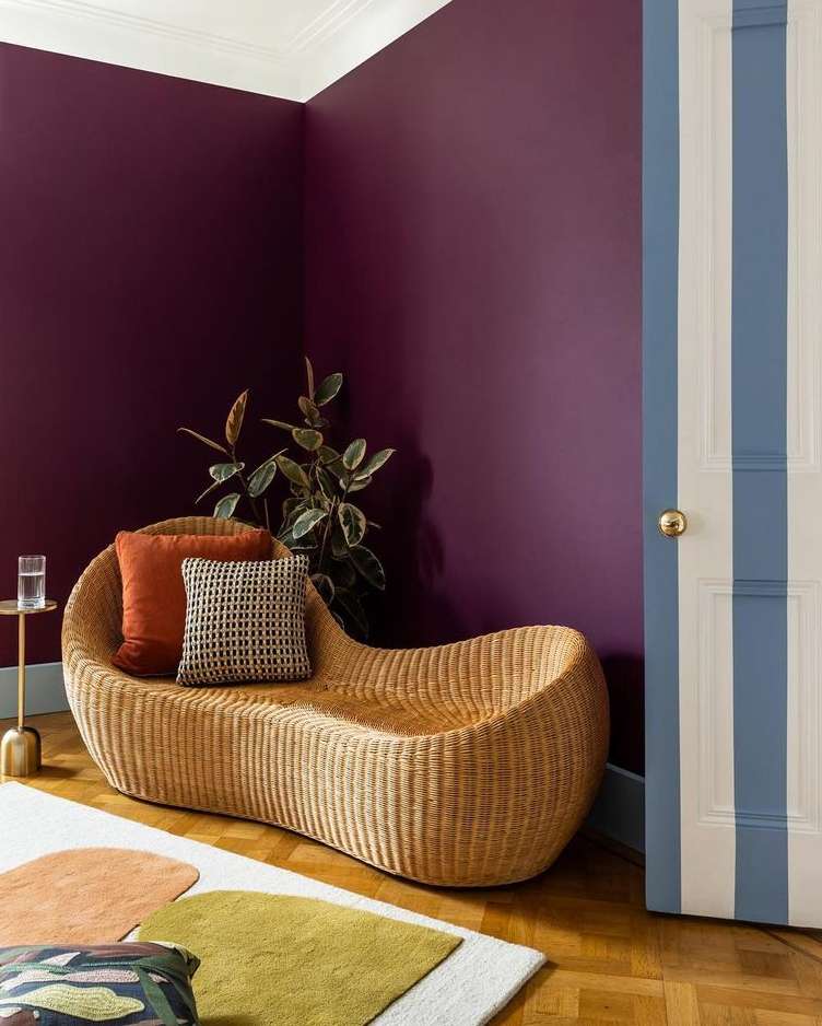 Interior shot showcasing a wicker chaise lounge with pillows, a side table with glass, and colorful geometric rugs against a plum wall.