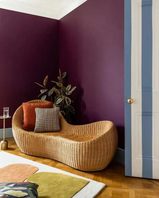 Interior shot showcasing a wicker chaise lounge with pillows, a side table with glass, and colorful geometric rugs against a plum wall.