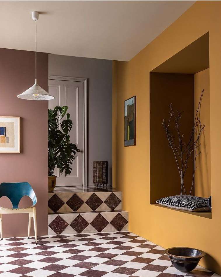 Indoor home interior featuring walls in rose, grey, and gold, with checkered floors, a modern chair, and potted plants near the doorway.