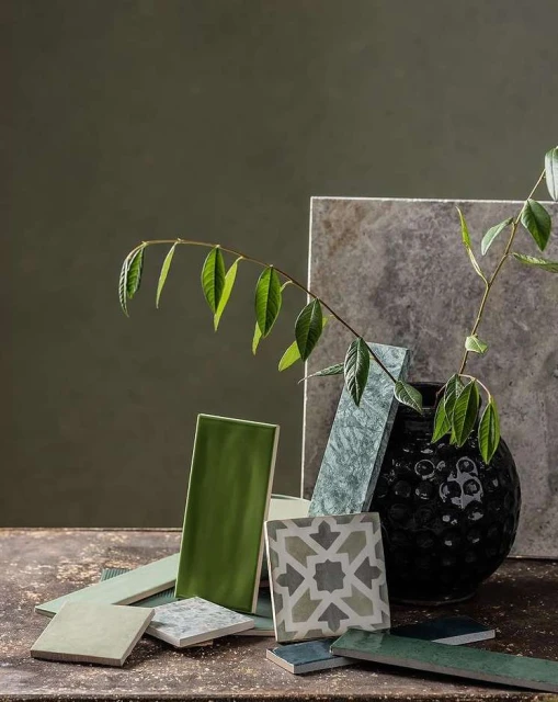 Still life composition featuring a collection of colorful tiles arranged with a plant stem and a dark vase on a textured surface.